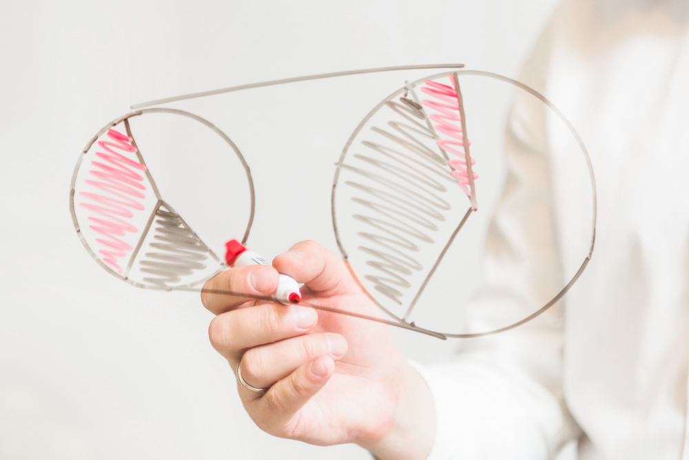Hand holding a marker and a wireframe model of eyeglasses with red and gray shading on the lenses, against a light background.