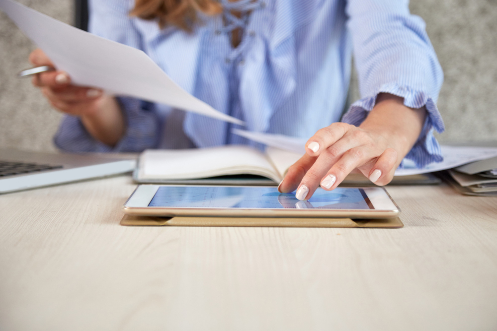 Person in a blue shirt uses a tablet on a desk, holding papers, with a notebook and laptop nearby.