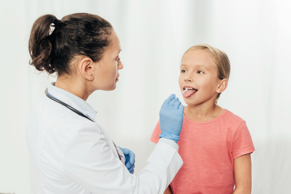 Doctor in white coat examines a girl’s throat with a tongue depressor. The girl, wearing a pink shirt, sits calmly with her tongue out.