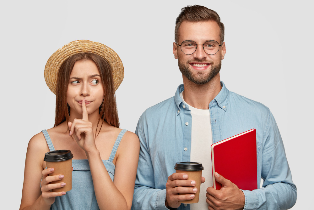 A woman in a straw hat gestures for silence while holding a coffee cup. A man beside her smiles, holding a coffee cup and a red folder.